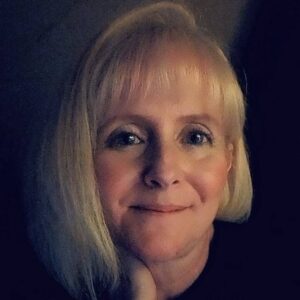 Author headshot of author Jamie Targaet, a blonde woman smiling softly in low light against a dark background.