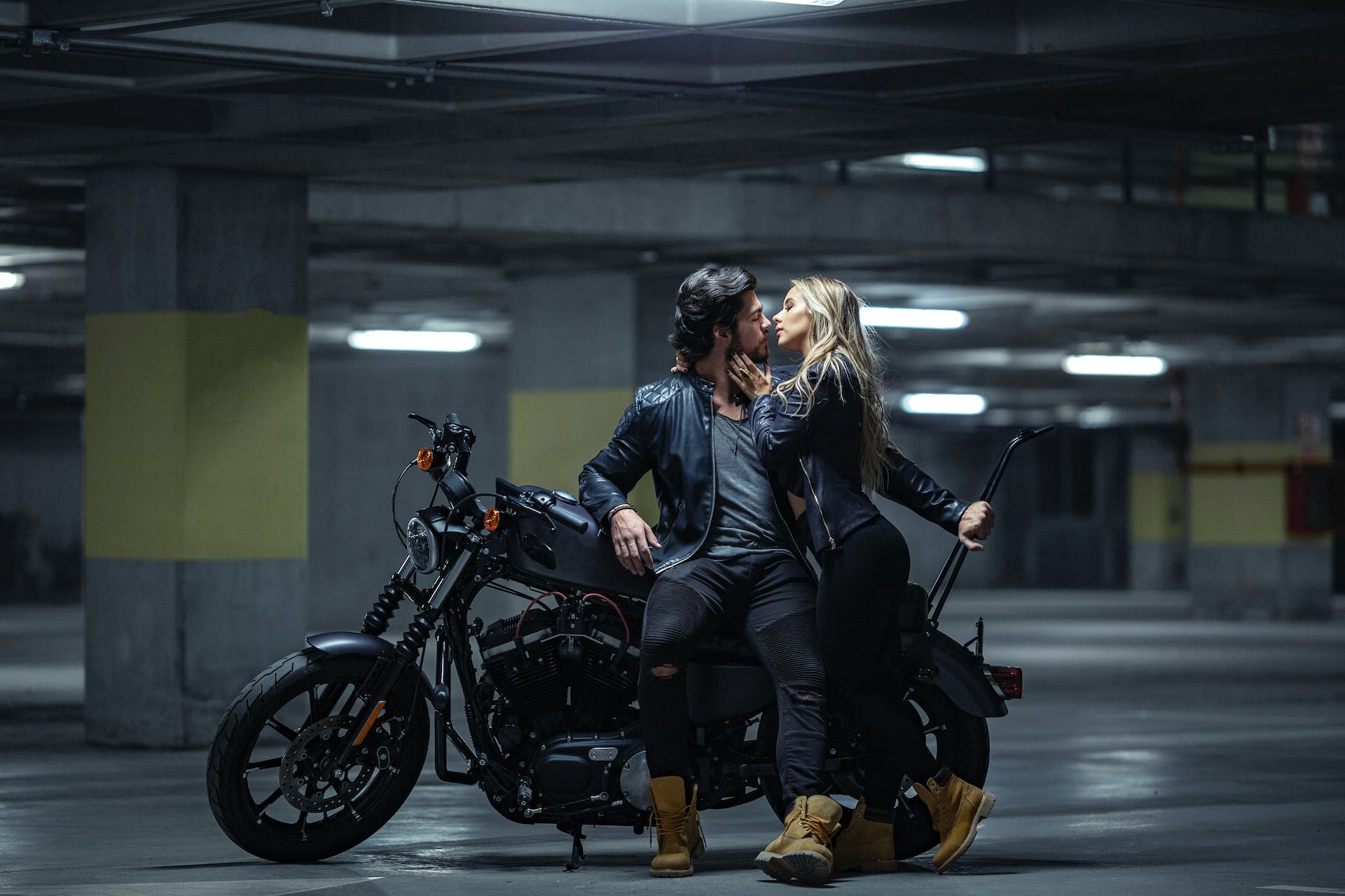 Romantic couple sharing an intimate moment while leaning against a black motorcycle in an underground parking garage